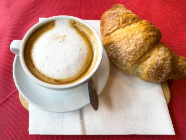 Kırmızı masa örtüsü üzerinde kruvasan olan kapuçino: sabah kahve molası sahnesi.