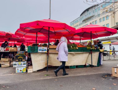 Zagreb, Hırvatistan - 14 Aralık 2025: Beyaz önlüklü kadın kırmızı şemsiyeler ve yerel satıcıların taze ürünleriyle dolu açık hava pazarında dolaşıyor