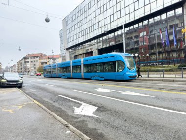 Zagreb, Hırvatistan - 14 Aralık 2025: Çağdaş binalar ve kentsel geçişi gösteren bulutlu bir atmosferle çevrili mavi tramvay şehir caddesinde ilerliyor