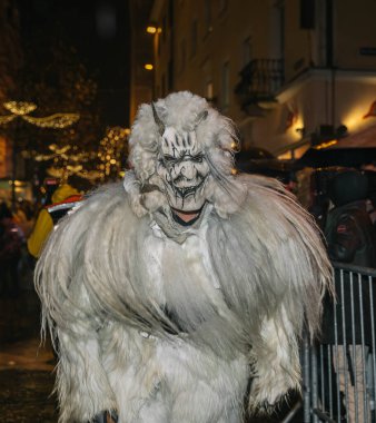 Villach, Avusturya - 01 Aralık 2023: Krampuslauf döneminde beyaz kürklü Krampus kostümlü bir adam, bayram gelenekleri ve kültürel kutlamaları somutlaştırarak festival caddesinde yürüyor