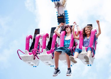 Villach, Avusturya - 29 Temmuz 2025: İki neşeli kız açık mavi gökyüzüne karşı parlak pembe koltuklu heyecan verici bir lunapark gezisi yaşıyor