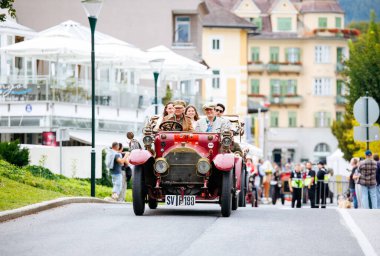 Velden, Avusturya - 30 Ağustos 2025: Klasik arabalar ve coşkulu kalabalıklarla dolu festival caddesinde gülümseyen yolcuların bulunduğu klasik kırmızı araba
