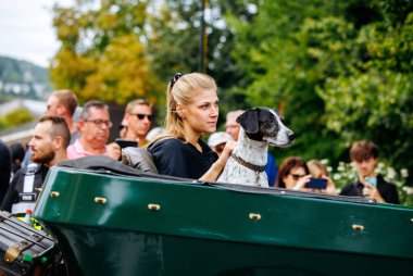 Velden, Avusturya - 30 Ağustos 2025: Genç sarışın kadın, festival katılımcılarıyla çevrili, köpeğiyle klasik bir teknede oturuyor.