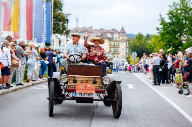 Velden, Avusturya - 30 Ağustos 2025: Klasik giysili neşeli çift festival sırasında festivalin keyfini çıkaran coşkulu kalabalıkla çevrili klasik eski zaman arabasını sürüyor