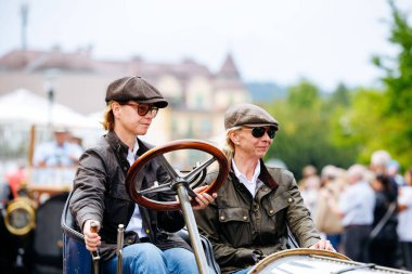 Velden, Avusturya - 30 Ağustos 2025: Şık şapkalı iki kadın, coşkulu kalabalık ve klasik araçlarla çevrili bir Oldtimer Festivali 'nde klasik bir araba kullanıyor