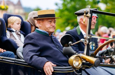 Velden, Avusturya - 30 Ağustos 2025: Geleneksel kıyafetler giymiş yaşlı bir adam, Oldtimer Festivali 'nde klasik arabasında, coşkulu izleyiciler ve klasik araçlarla çevrili oturuyor