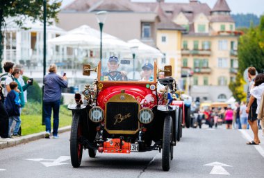 Velden, Avusturya - 30 Ağustos 2025: İki şoförlü klasik kırmızı araba coşkulu izleyicilerle dolu hareketli bir festival caddesinden geçiyor