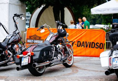 Velden, Avusturya - 30 Ağustos 2025: Harley-Davidson bisikleti, açık hava etkinliğinin keyfini çıkaran coşkulu kalabalıkla çevrili turuncu bir pankartın yanında belirgin bir şekilde sergileniyor