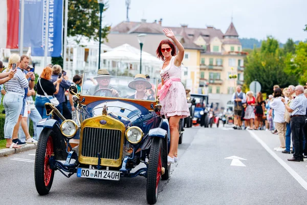 Velden, Avusturya - 30 Ağustos 2025: Klasik klasik araba geçit töreni arka planda heyecanlı kalabalıkla birlikte antik bir araçtan el sallayan neşeli bir kadın sergileniyor