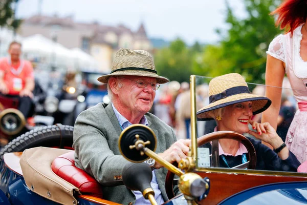 Velden, Avusturya - 30 Ağustos 2025: Yaşlı çift, klasik araçlar ve neşeli katılımcılarla çevrili Oldtimer Festivali 'nde klasik araba sürerken gülümsüyor