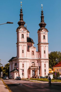 Villach, Avusturya - 14 Ağustos 2025: Barok tarzı pembe kilise, iki uzun kuleli ve altın yıldızlı, güneşli bir caddede, araçları ve yakındaki bir bisikletçiyle