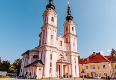 Villach, Avusturya - 14 Ağustos 2025: Güneşli bir meydanda, renkli binalar ve yeşilliklerle çevrili iki uzun kuleli tarihi pembe kilise