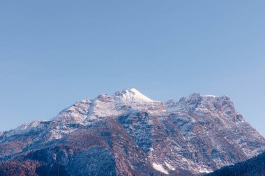 Dolomite dağının karla kaplı zirvesi açık mavi gökyüzüne karşı yükseliyor, engebeli uçurumları ve Alp bölgesinin doğal güzelliklerini vurguluyor.