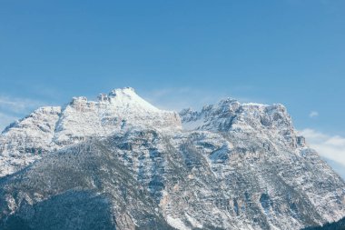 Dolomitlerin karla kaplı dağları parlak mavi bir gökyüzüne karşı görkemli bir şekilde yükselirler. Cortina 'nın alçak yamaçlarında yoğun yeşil ormanlar görülür.