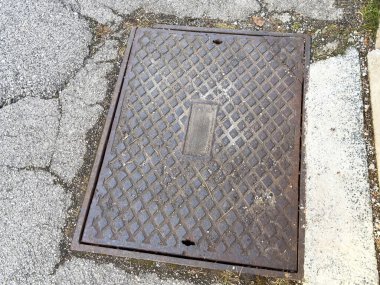 Çatlak kaldırımda desenli tasarımı olan paslı kare rögar kapağı..