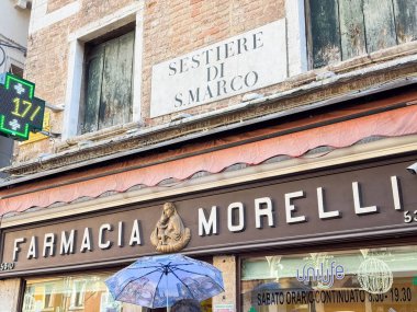 Venedik, İtalya - 17 Şubat 2026: Farmacia Morelli 'nin mağazası Sestiere di S. Marco işareti, dekoratif figür ve şemsiye tutan birini gösteriyor.