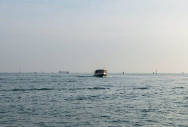 Venedik, İtalya - 7 Şubat 2026: Uzak yelkenlileri ve berrak gökyüzü ile sakin sularda hareket eden küçük tekne, barışçıl bir deniz ortamı sergiliyor.