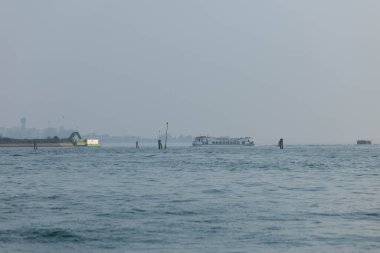 Venedik, İtalya - 07 Şubat 2026: Tekne sisli bir siluet ve kıyı şeridi boyunca uzak yapılar ile huzurlu bir kıyı sahnesi oluşturarak sakin sularda ilerliyor