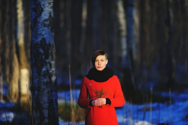 Retrato de primavera temprana de linda chica joven atractiva seria con bufanda de calor de pelo oscuro y chaqueta roja mirando a la cámara y de pie en el bosque salvaje con una ramita en las manos. Exterior — Foto de Stock