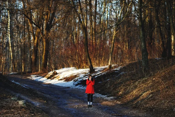 La chica de abrigo rojo estaba congelada en medio de un bosque de primavera. Ella está tratando de calentar sus manos. — Foto de Stock