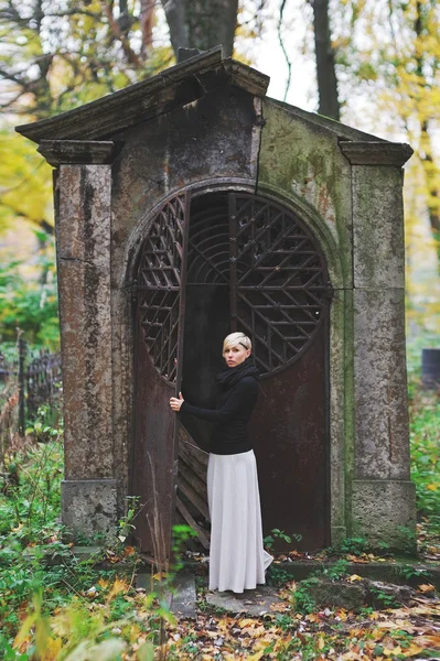 Joven rubia con estilo en un jersey negro y falda blanca larga se encuentra cerca del monumento en el cementerio, sosteniendo sus manos detrás de puertas de hierro mira a la cámara . — Foto de Stock