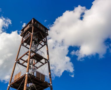 Uzun, ahşap bir ateş gözetleme kulesi, tüylü, beyaz bulutlu, mavi bir gökyüzüne karşı duruyor. Yapı metal ve ahşaptan yapılmıştır, kırsal bir tasarım sergiler..
