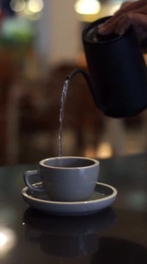 Sıcak su kazan boynundaki çaydanlıktan seramik bardağa sıcak bir iç ortam ve yumuşak bokeh ile akıyor. İçecek, kafe yaşam tarzı, demleme ve aşçılık temaları için ideal.