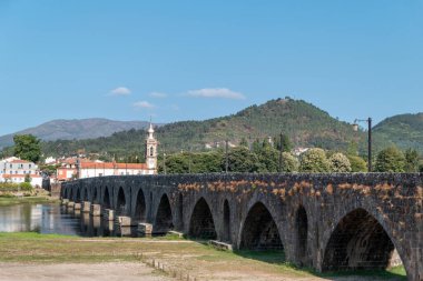 Lima Nehri üzerindeki Ponte de Lima Ortaçağ ve Roma köprüsü, ana kilise ve çan kulesi ve mavi gökyüzünün altında yeşil Minho dağları.
