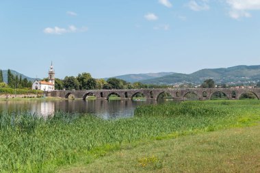 Panoramic view of the Medieval and Roman Bridge of Ponte de Lima over the Lima River, highlighting the Church of Santo Antonio da Torre Velha, flanked by trees and framed by riverside vegetation in Portugal