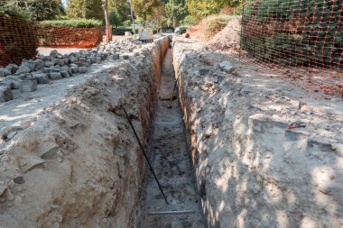 A deep, narrow trench dug in an urban area for sanitation infrastructure work, with soil removed and piled on the sides, and some tools scattered throughout the trench
