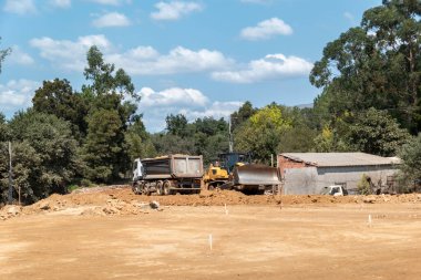 Toprağı boşaltan bir çöp kamyonu ve bir buldozer güneşli bir günde arka planda yoğun bitki örtüsü bulunan bir otoyol inşaatında toprak taşıma işi yapıyorlar.