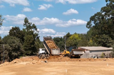 Toprağı boşaltan bir kamyon ve bir buldozer, dağılmış bulutlarla dolu mavi bir gökyüzünün altında yemyeşil bir bitki örtüsüyle çevrili bir otoyol inşaatında toprak çalışmaları yapıyorlar.