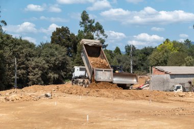 Buldozer yollarda çalışırken bir kamyon toprağı boşaltıyor. Mavi gökyüzünün altında bulutlarla benekli bitki örtüsüyle çevrili.