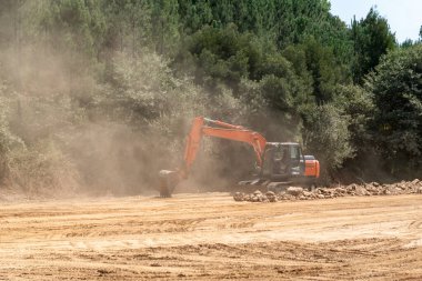 Otoyol inşaatında yer değiştiren bir kazıcı ağaçlık bir alanda ağır bir toz bulutu yaratıyor.