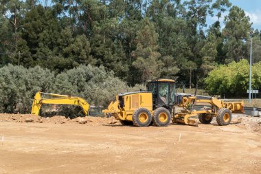 Otoyol inşaatı için arazi hazırlığı zemini dengeleyen bir motor greyderi ve çevresi bitki örtüsüyle çevrili bir inşaat alanında çalışan bir kazıcı ile.