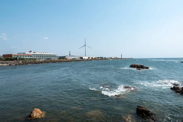Viana do Castelo 'daki kıyı manzarası, küçük kaya oluşumları üzerinde dalgalı denizler, modern endüstriyel binalar ve Portekiz' in arka planında bir rüzgar türbini barındırıyor.