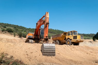 Hidrolik kazıcı ve çöp kamyonu tamamen toprak taşıma operasyonunda, mavi gökyüzünün altındaki konut binalarının gelişimi için zemin hazırlıyor.