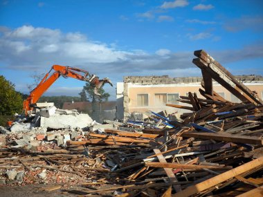 Kazıcı, engin bir moloz yığını üzerine yerleştirilmiş, hasarlı ahşap, tuğla ve beton parçalarından oluşan bir binanın yıkımında çalışıyor.
