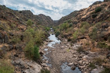 Faia Brava Doğal Parkı 'ndaki Coa Nehri' nin ıssız manzarası, Portekiz 'deki Dostluk Köprüsü' nün yanındaki bulutlu gökyüzünün altındaki kayalık, kuru bitki örtüsü yamaçlarıyla çevrili taşlı nehir yatağı.