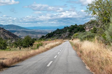 Parçalı bulutlu bir gökyüzünün altında dağlık bir araziyi kesen dolambaçlı asfalt bir yol.