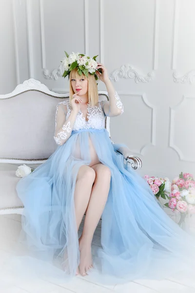 Beautiful pregnant woman in light blue dress with flower diadem in white room