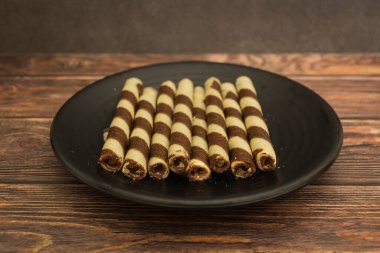 Kue Semprong olarak bilinen gevrek ve lezzetli Endonezya gofretleri, basit bir siyah plaka seti üzerinde sade, koyu kahverengi ahşap arka plana karşı düzenlenmiştir..