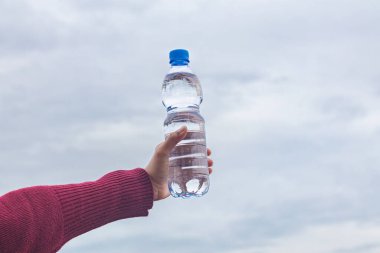 Kadının eli arka planda bulutlu gökyüzü olan bir su şişesini tutuyor.