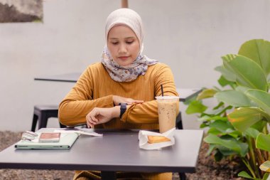 Asyalı Müslüman bir kadın kahve dükkanında otururken saatine bakıyor.