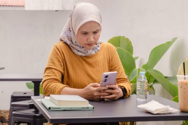 Asyalı Müslüman bir kadın açık bir kafede akıllı telefonuna mesaj yazıyor.