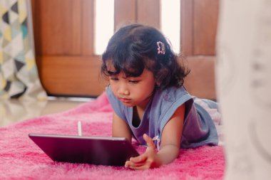 Küçük bir Asyalı kız yerde dinlenirken öğrenmek ve oynamak için tablet ve kalem kullanıyor.