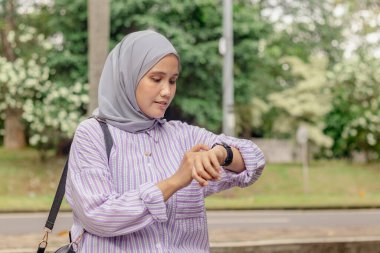 Genç bir Asyalı Müslüman kadın parkta yürürken saatine bakıyor.