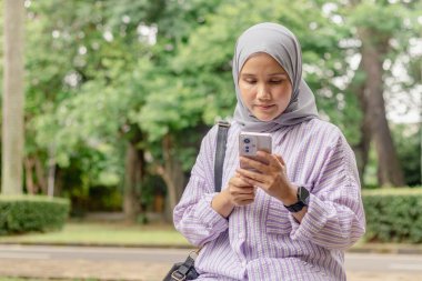 Genç bir Asyalı Müslüman kadın dışarıda kadın çantası taşırken akıllı telefonunu kullanıyor.
