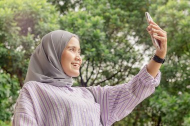Mutlu bir Asyalı genç Müslüman kadın video görüşmesi yapıyor ya da dışarıda cep telefonuyla selfie çekiyor.