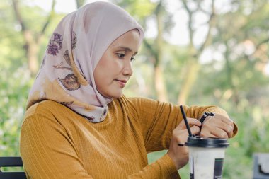 Endonezyalı Müslüman bir kadın parkta otururken saatine bakıyor.